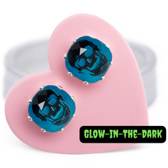 A pair of Glow in the dark earrings, featuring skulls on a Bright Aqua background, carded on a pink heart.