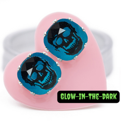 Blue stud earrings with a skeleton in the center, carded on a pink heart, in the light.